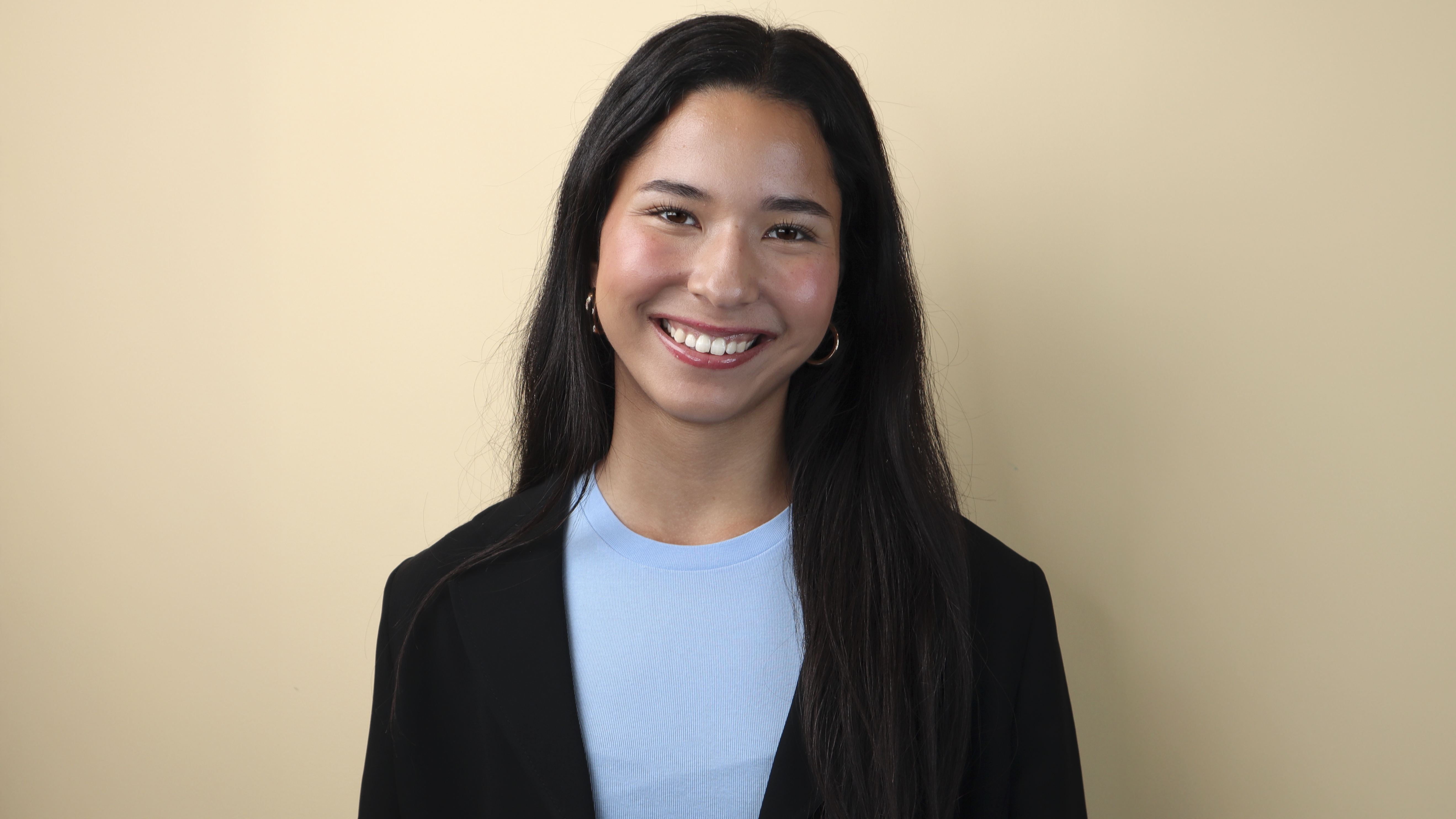 Anastasia Chostner smiles in front of a beige background while wearing a black blazer over a light blue shirt. Anastasia Chostner smiles in front of a beige background while wearing a black blazer over a light blue shirt.